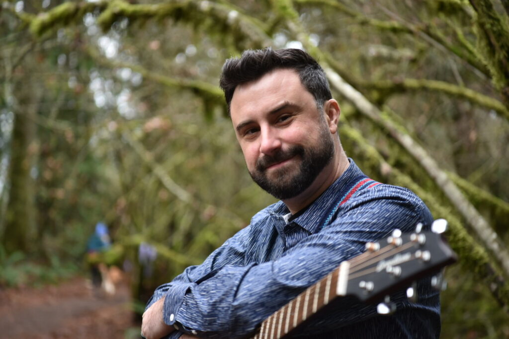 Bobby Jo Valentine, a white man with dark short hair and a beard wearing a long-sleeve blue shirt, leans on a guitar while sitting on the edge of a trail in the woods.