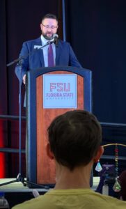 Donald Post, a white man with glasses and short brown hair and a beard, stands at a podium wearing a blue suit and red patterned tie. The placard on the front of the podium says FSU Florida State University. 