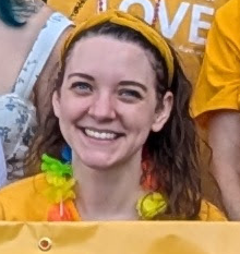 Emily Brier, a young white woman with long brown hair, smiles at the camera. She is wearing a yellow Side with Love shirt, a vibrant Hawaiian lei, and a yellow headband.