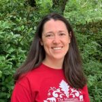 This is a photo of Anna Martin, a white woman with dark brown hair, just past her shoulders. She is wearing a red t-shirt that says "The Mountain" on it, and she is smiling as she stands in front of a very green rhododendron bush.
